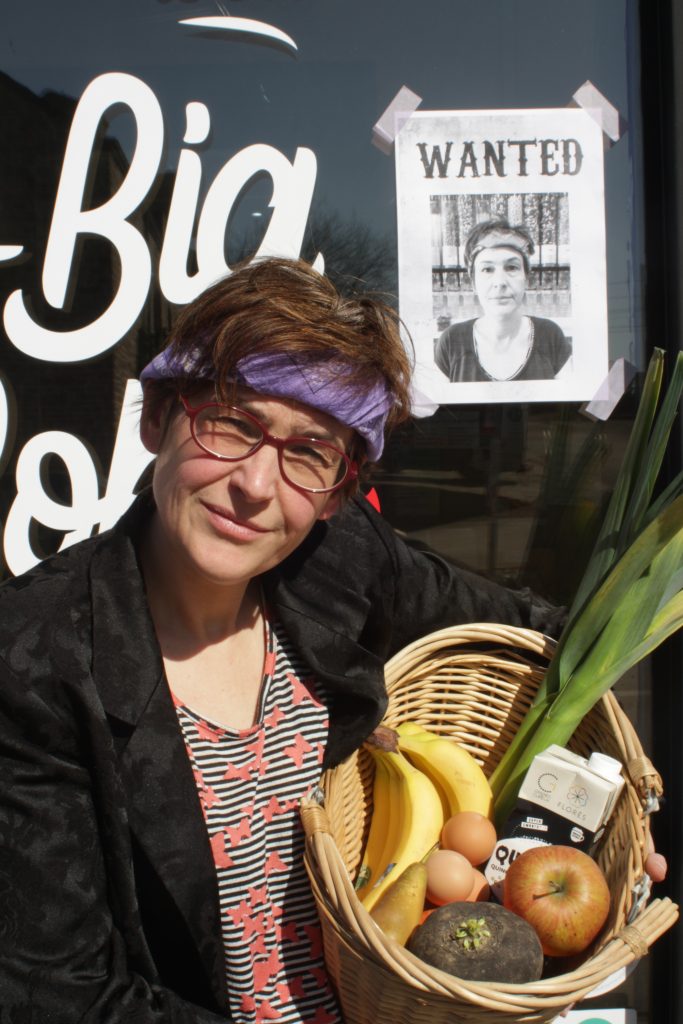 Odile pose avec un panier de légumes devant son magasin. Elle est coifée d'un bandana et a la tête un peu inclinée comme une vedette des années 80.